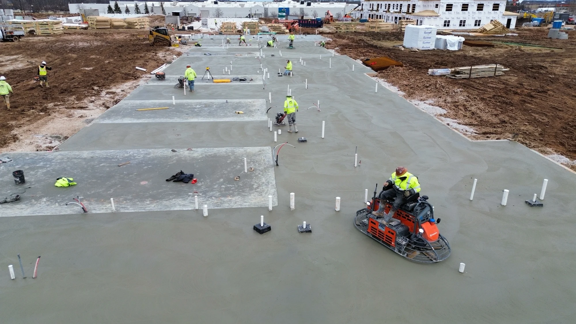 Construction workers in yellow safety gear smoothing concrete foundation at building site.