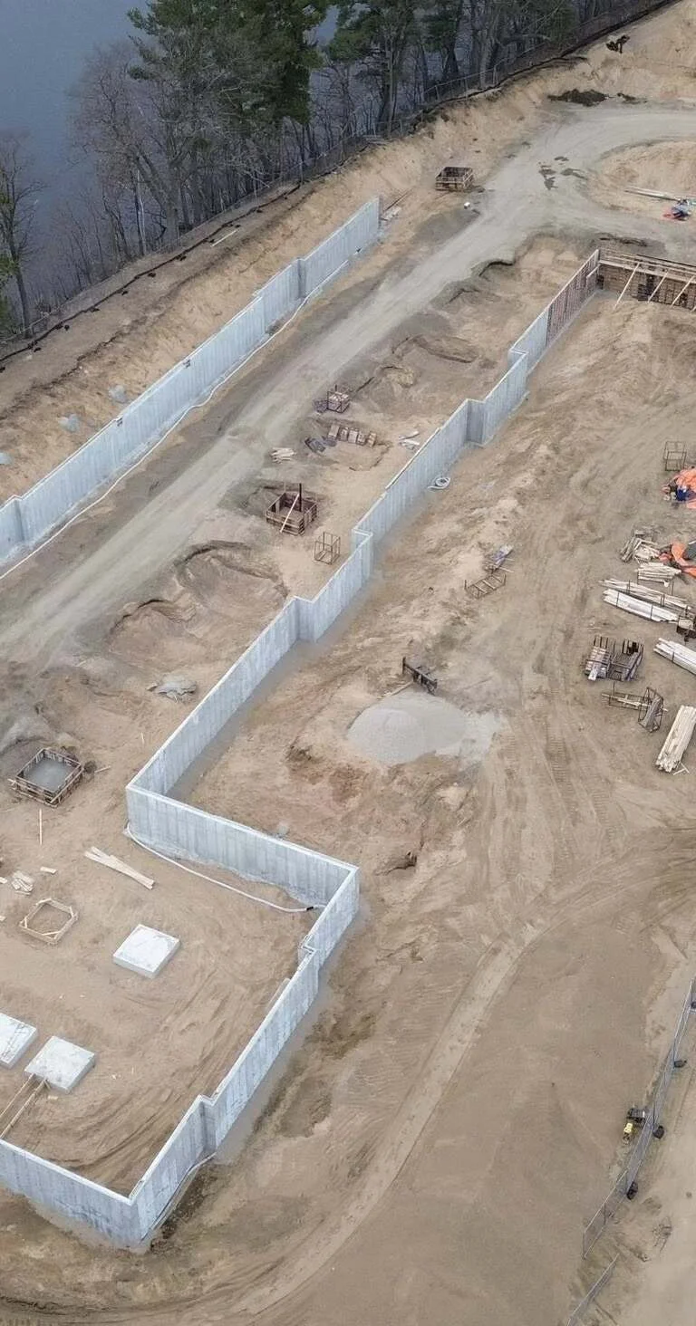 Aerial view of a construction site with concrete retaining walls, dirt ground, construction materials, and surrounding trees.