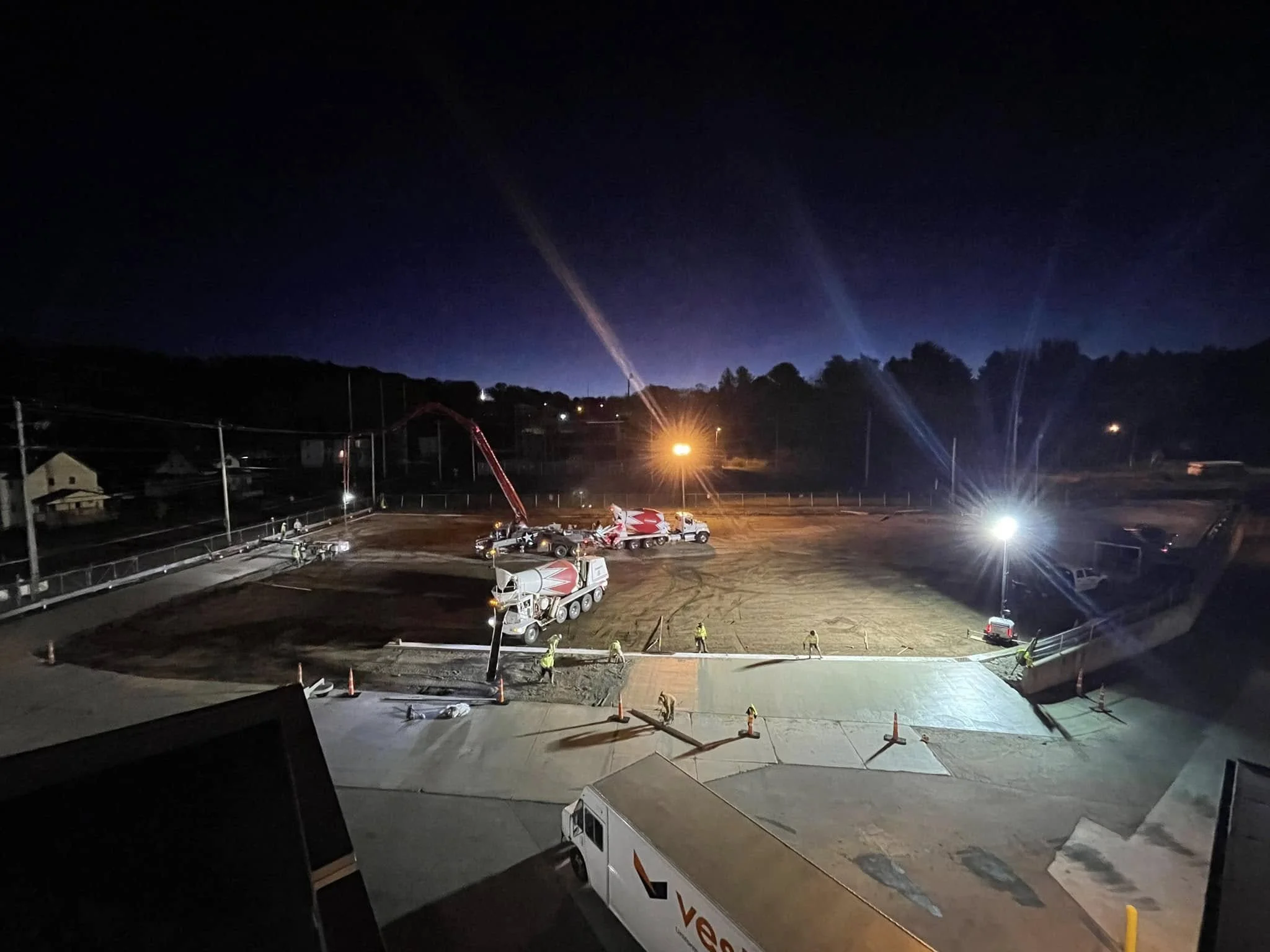 Nighttime construction site with cement trucks, workers, and bright floodlights illuminating the area.