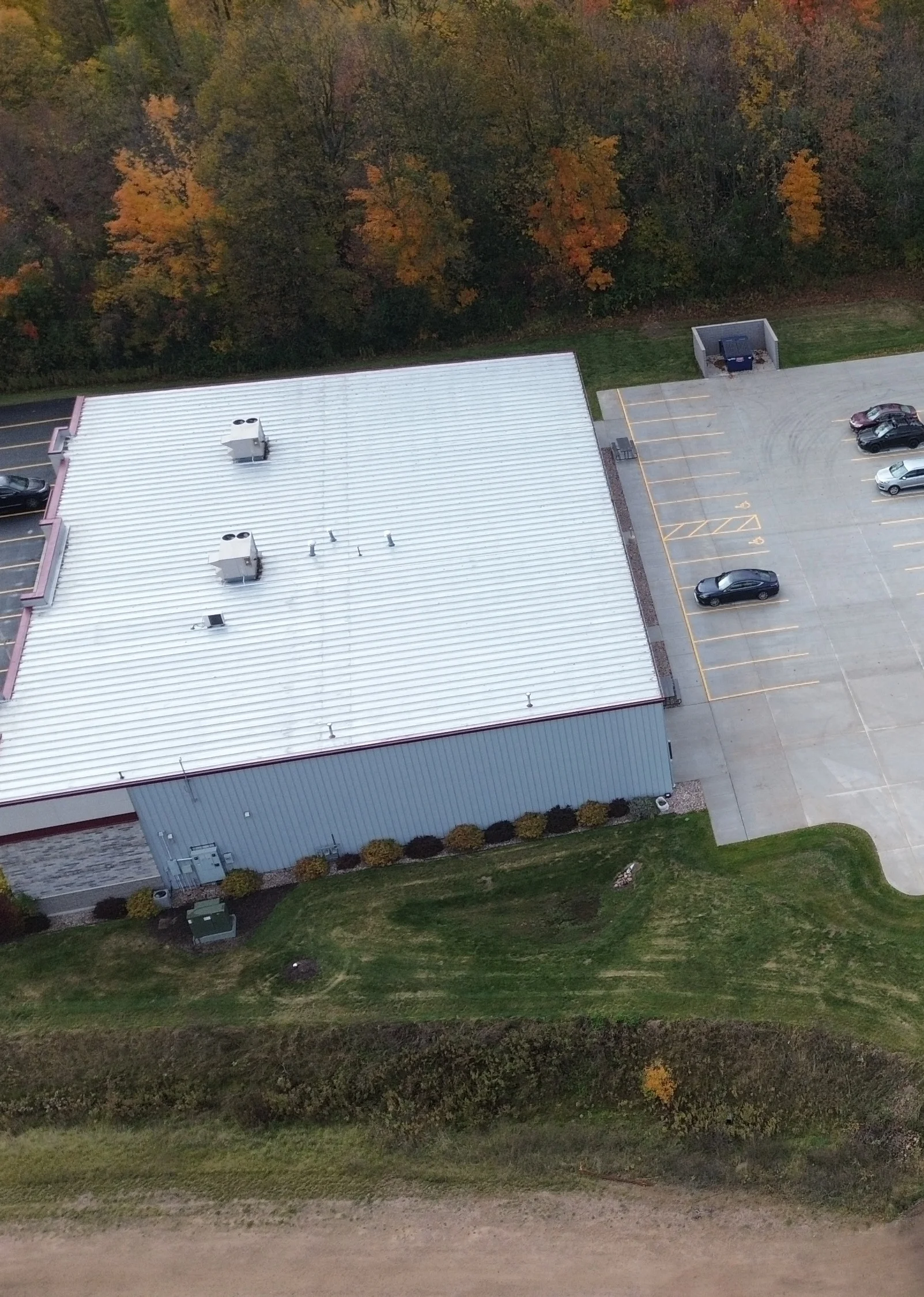 Aerial view of a white-roofed industrial building, adjacent parking lot with cars, and autumnal trees.