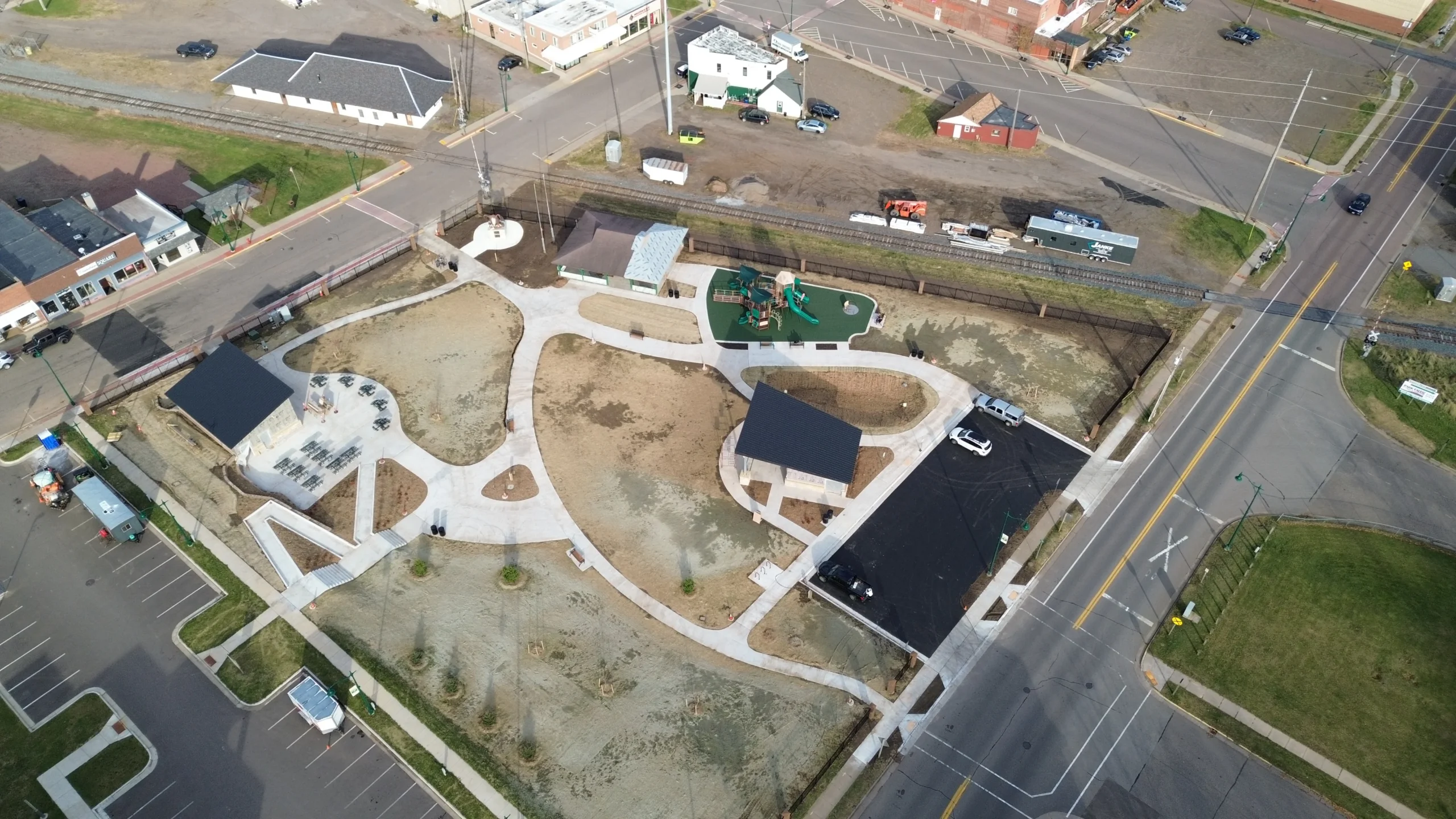 Aerial view of a small urban park with playground, seating areas, pathways, adjacent streets, and nearby buildings.