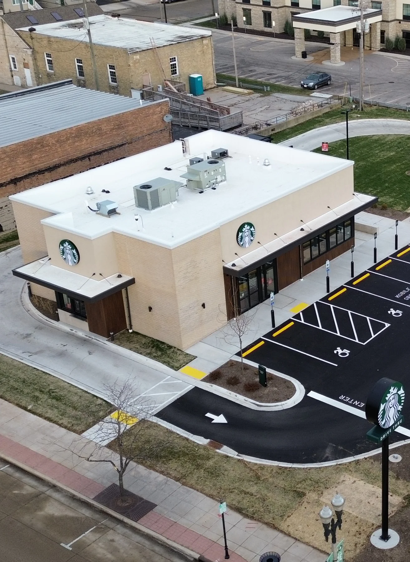 Aerial view of a beige Starbucks building with drive-thru, marked parking spaces, and adjacent brick buildings.