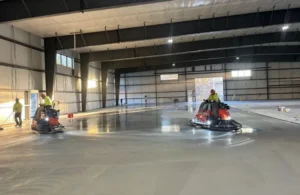 Workers operating floor polishing machines on a large concrete surface inside a spacious industrial warehouse.