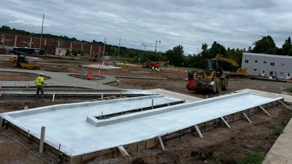 Construction site with workers, heavy machinery, and freshly poured concrete pathways under a cloudy sky.