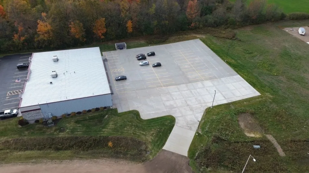 Aerial view of a building with a metal roof, adjacent large parking lot with few cars, and surrounding greenery with autumn-colored trees.